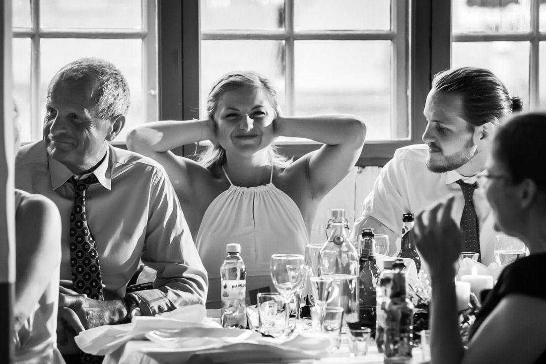 Bruden ler under talet Bruden ler med händerna i håret mellan pappa och brudgum under ett tal i svartvitt – bröllopsfotograf Skåne