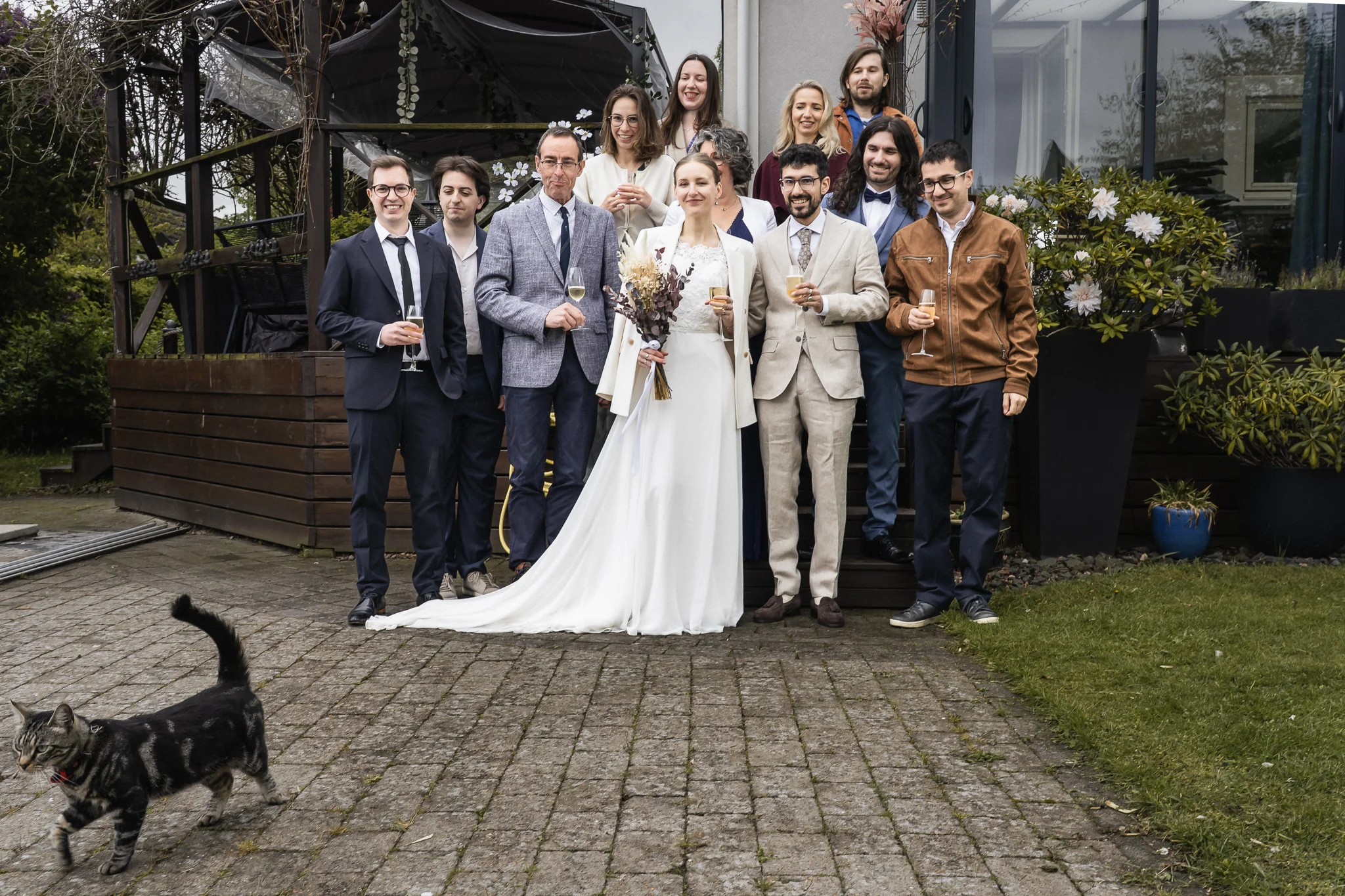 Gruppbild med hela sällskapet Gruppbild på brudpar och gäster utanför hemmet i Malmö, med en katt i förgrunden – bröllopsfotograf Malmö