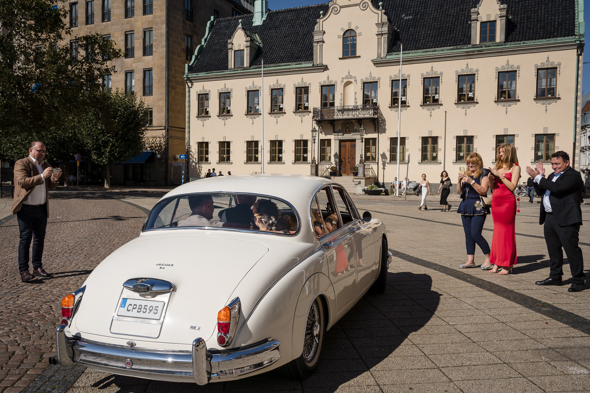 Bilen lämnar torget Vit Jaguar kör över torget framför Malmö Rådhus med vinkande gäster – bröllopsfotograf Malmö